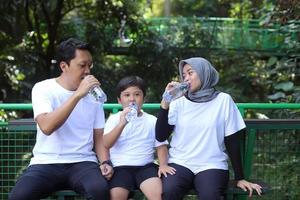 Glückliche muslimische Familie, die eine Flasche mit Wasser hält und trinkt, während sie sich nach dem Sporttraining im Freien im öffentlichen Park ausruht. foto