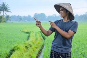 attraktiver, fröhlicher junger asiatischer Bauer, der zur Seite lächelt und mit dem Finger auf die Seite des Reisfeldes mit Kopierraum zeigt. modernes Landwirtschaftskonzept. foto