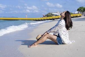 Besorgte Frau, die mit einem Strohhut auf den Knien am Strand sitzt Tropischer Strand am Meer bei sonniger Tageszeit. Einsames Mädchen, das allein am Meer sitzt, sich entspannt und nachdenkt. Konzeptfotografie menschlicher Emotionen foto