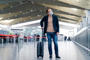 Ganzkörperaufnahme eines Passanten mit Koffer auf dem internationalen Flughafen trägt eine schützende Gesichtsmaske, um eine Coronavirus-Infektion zu vermeiden. Männchen mit Gepäck, das ins Ausland reisen wird. epidemie, gesundheitswesen foto