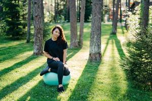 fitte brünette junge frau ruht sich aus, nachdem sie übungen am fitnessball gemacht hat, führt aktive sportliche lebensstilposen in städtischer umgebung auf grünem gras gegen bäume mit sonnenschein turnt. foto