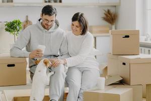 positives junges Ehepaar spielt mit Hund, sitzt auf Kartons, trinkt Kaffee zum Mitnehmen, macht Pause am Umzugstag und packt Sachen aus, trägt Freizeitkleidung, genießt das Leben in der neuen Wohnung. foto