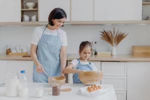 Foto von brünetter Mutter und kleinem Kind machen Plätzchenteig, verquirlen Zutaten in einer Schüssel, tragen Schürzen, Mutter freut sich, einen kleinen Helfer in der Küche zu haben. zusammen kochen, familien- und heimkonzept