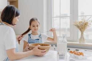 Aufnahme eines kleinen Kinderhelfers schlägt Sahne mit Schneebesen auf, lernt backen, hört nützliche Ratschläge von Mutter, die ihren Pferdeschwanz hält, sitzt zusammen am weißen Küchentisch. Familie zusammen kochen foto