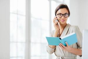 foto einer attraktiven frau mit dunklen haaren, führt ein telefongespräch, willigt ein, sich mit einem kollegen zu treffen, hält ein geöffnetes tagebuch in der hand, trägt eine brille und einen formellen anzug, posiert vor weißem hintergrund, plant den tagesplan