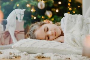 nahaufnahme eines attraktiven kleinen kindes liegt auf dem boden, schläft in der nähe von neujahr oder weihnachtsbaum, wärmt mit weißem strickpullover, hat angenehme träume am abend. kinder, winterferien, restkonzept foto