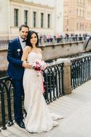 hübscher bärtiger bräutigam umarmt seine braut im weißen kleid mit blumenstrauß, steht zusammen auf der brücke, hat gute laune nach der registrierung ihrer ehe. gutes beziehungskonzept foto