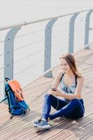 außenaufnahme einer angenehm aussehenden entspannten frau sitzt mit gekreuzten beinen, schaut positiv auf das smartphone, hört musik mit kopfhörern und genießt highspeed-internet. Reisender macht nach langem Spaziergang Pause foto