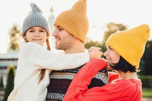 Menschen und Beziehungskonzept. familie unvergessliche zeit miteinander verbringen, sich umarmen, trendige strickmützen tragen. entzückendes kleines Mädchen mit Zöpfen umarmt ihren Vater, schaut glücklich in die Kamera foto