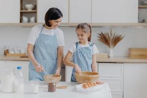 innenansicht der glücklichen mutter und tochter bereiten gemeinsam ein leckeres abendessen zu, stehen in schürzen neben dem küchentisch nebeneinander, lächeln und verquirlen eier in der schüssel. menschen, familie und kulinarisches konzept foto