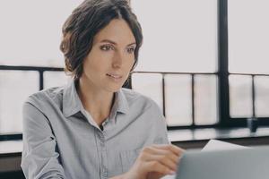 junge weibliche Büroangestellte, die am Arbeitsplatz sitzen und online am Laptop arbeiten, foto