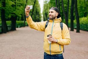 Isoliertes Porträt eines Hipster-Typen in modischer Kleidung, der allein im Park ruht und gutes Wetter und frische Luft bewundert. bärtiger mann, der selfie im park macht und kaffee zum mitnehmen trinkt, der im park steht foto