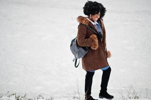 lockiges haar afroamerikanerin trägt schaffellmantel und handschuhe mit rucksack, der am wintertag gestellt wird. foto