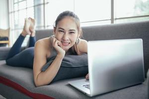 junge asiatische frau, die einen laptop verwendet, der zu hause die social-media-anwendung auf der website online überprüft, fühlt sich entspannt und genießt es, im wohnzimmer zu kommunizieren und zu diskutieren. foto