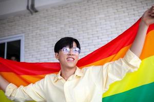 asiatischer schwuler, der zu hause lgbt-regenbogenfahne hält, lgbtq-konzept. foto