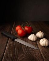 Knoblauchzwiebeln, Tomaten und ein Messer auf einem dunklen Holztisch mit Kopienraum oben foto