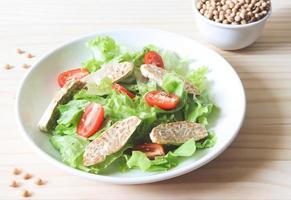 Tempeh- oder Tempe-Salat mit Tomaten und grünem Gemüse in weißer Platte und einer Schüssel Sojabohnen auf Holztisch. foto