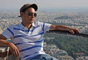 hübscher junger mann mit baseballmütze und sonnenbrille sitzt in der sonne mit der stadt athen im hintergrund foto