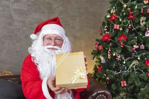 der weihnachtsmann hält eine weihnachtsgeschenkbox mit einem vollständig geschmückten chrsitmas-baum für die jahreszeitfeier und ein frohes neues jahr foto
