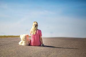 Mädchen im karierten Anzug sitzt mit Teddybär auf Asphaltstraße. konzept der einsamkeit, die auf glück wartet foto