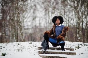 lockiges haar afroamerikanerin trägt schaffellmantel und handschuhe mit rucksack, der am wintertag gestellt wird. foto