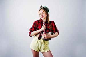 junge lustige Hausfrau in kariertem Hemd und gelben Shorts Pin-up-Stil mit Topf und Küchenlöffel isoliert auf weißem Hintergrund. foto