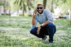 stylischer afroamerikanischer junge auf grauem pullover und schwarzer sonnenbrille posierte im park und sprach am telefon. Modischer Schwarzer. foto