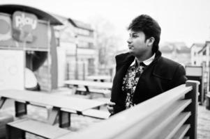 Attraktiver und gutaussehender, stilvoller indischer Mann trägt einen schwarzen Mantel, der in seiner Arbeitspause auf der Terrasse des modernen Lounge-Cafés steht. foto