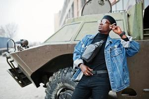 afroamerikanischer mann in jeansjacke, baskenmütze und brille, rauchende zigarre und posierte gegen btr militärisches gepanzertes fahrzeug. foto