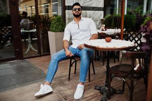 Stilvolles großes arabisches Mannmodell in weißem Hemd, Jeans und Sonnenbrille posierte auf der Straße der Stadt. Bart attraktiver arabischer Typ, der auf einem Café im Freien sitzt. foto