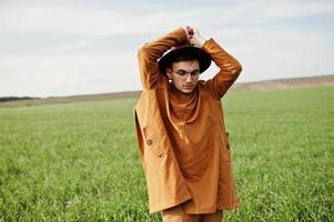 stylischer mann mit brille, brauner jacke und hut posierte auf der grünen wiese. foto