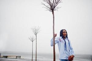 Stilvolles afrikanisch-amerikanisches Mädchen mit Dreadlocks, das Handy zur Hand hält, im Freien auf Pier gegen zugefrorenen See bei Schneewetter. foto