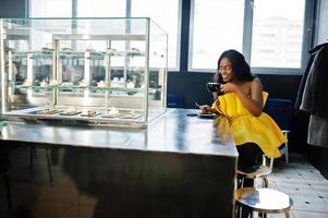 Stilvolle afroamerikanische Frau im gelben Kleid posierte im Café mit einer Tasse Cappuccino. foto