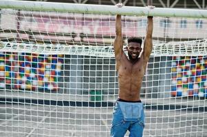 hübscher sexy afroamerikanischer muskulöser Mann mit nacktem Oberkörper in Jeans-Overalls posiert auf grünem Gras gegen Fußballtore des Stadionfeldes. modisches Porträt eines schwarzen Mannes. foto