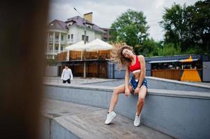 sexy lockiges Model-Mädchen in rotem Oberteil, Jeans-Denim-Shorts, Brille und Turnschuhen posiert im Skatepark. foto