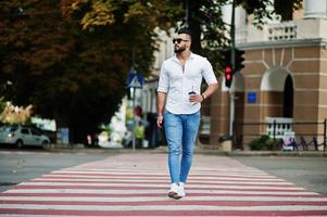 Stilvolles großes arabisches Mannmodell in weißem Hemd, Jeans und Sonnenbrille posierte auf der Straße der Stadt. bart attraktiver arabischer kerl mit tasse kaffee, der auf fußgängerüberweg oder zebrastreifen geht. foto