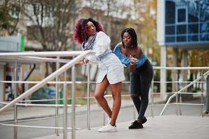 zwei attraktive afroamerikanische frauen posierten in der nähe von geländern gegen ein modernes mehrstöckiges gebäude. aufs Gesäß schlagen. foto