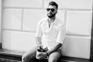 Stilvolles großes arabisches Mannmodell in weißem Hemd, Jeans und Sonnenbrille posiert auf der Straße der Stadt und hält das Handy zur Hand. bart arabischer kerl mit einer tasse kaffee sitzen und sich ausruhen. foto