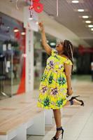 süßes afroamerikanisches mädchen mit kleiner höhe und dreadlocks, trägt ein farbiges gelbes kleid und zeigt den finger auf dem rabattschild im einkaufszentrum. foto