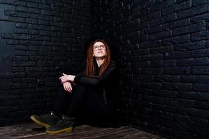 Studio-Shooting von Mädchen in Schwarz mit Dreads, Brille und Hut auf Backsteinhintergrund. foto