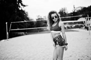 sexy lockiges Model-Mädchen in rotem Top, Jeans-Denim-Shorts, Tasse und Sonnenbrille am Beach-Volleyball-Feld. foto