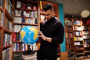 großer, intelligenter arabischer student, trägt einen violetten rollkragenpullover und eine brille, in der bibliothek, die erdkugel an den händen hält. foto