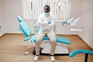 afroamerikanischer männlicher arzt in maske und brille mit verschränkten armen sitzt am zahnarztstuhl in der zahnklinik. foto