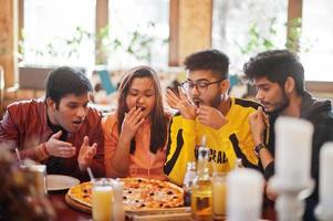 gruppe asiatischer freunde, die pizza während der party in der pizzeria essen. glückliche indische leute, die zusammen spaß haben, italienisches essen essen und auf der couch sitzen. schockierte und überraschte Gesichter. foto