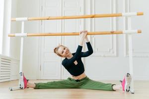 Kaukasische Ballerina spreizt die Beine seitwärts, zeigt Spagat, hält die Hände an Stangen, hat Ballettunterricht, trägt Sportkleidung und Turnschuhe, hat eine perfekte Figur, neigt den Kopf nach rechts, streckt sich. foto