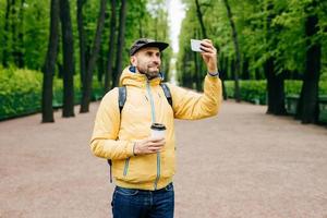 Outdoor-Porträt eines gutaussehenden Mannes mit dickem Bart, der einen gelben Anorak und Jeans trägt, die Rucksack, Kaffee und Smartphone halten, die Selfie gegen grüne Bäume machen. zufriedener tourist, der sich im park ausruht foto