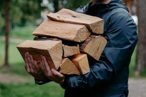 7/8-Aufnahme eines gesichtslosen Mannes in schwarzer Jacke trägt einen Haufen Brennholz-Posen vor verschwommenem Waldhintergrund. foto