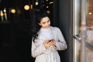 porträt einer modischen brünetten frau mit angenehmem aussehen, die eine schöne maniküre trägt und einen weißen regenmantel trägt, der ein smartphone in ihren händen hält und ein angenehmes lächeln hat, das froh ist, eine nachricht zu erhalten foto