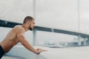 motivierter männlicher sportler macht push-up-übungsposen im freien an der brücke, konzentriert auf distanz, hat morgendliches training, nackter muskulöser oberkörper wärmt sich vor dem joggen auf. gesundes lebensstilkonzept. foto
