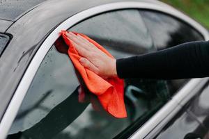 bemannt die hand mit lappen, der seitenfenster des schwarzen autos reinigt. professionelles autowaschkonzept. regelmäßiges Abwaschen. Abwischen von Wassertropfen am Fahrzeug foto